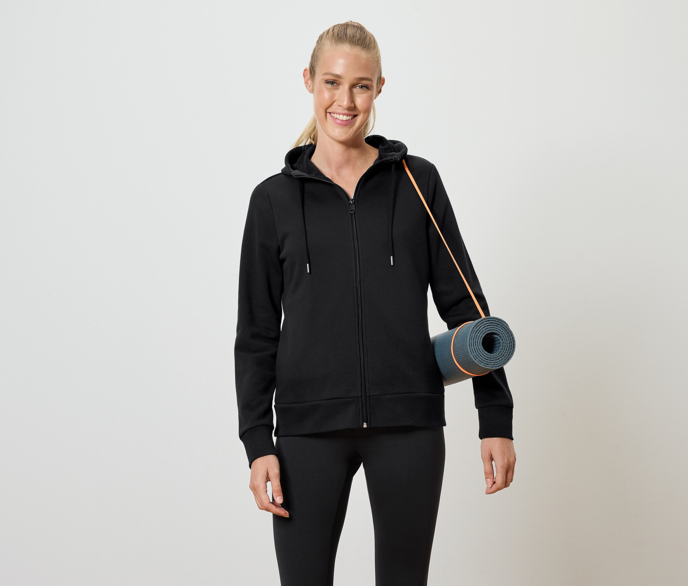 Femme en sweat à capuche noir zippé et leggings noirs tenant un tapis de yoga enroulé sous le bras.