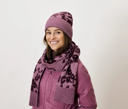 Une femme souriante porte une veste violette, une écharpe à motifs et un bonnet assorti.