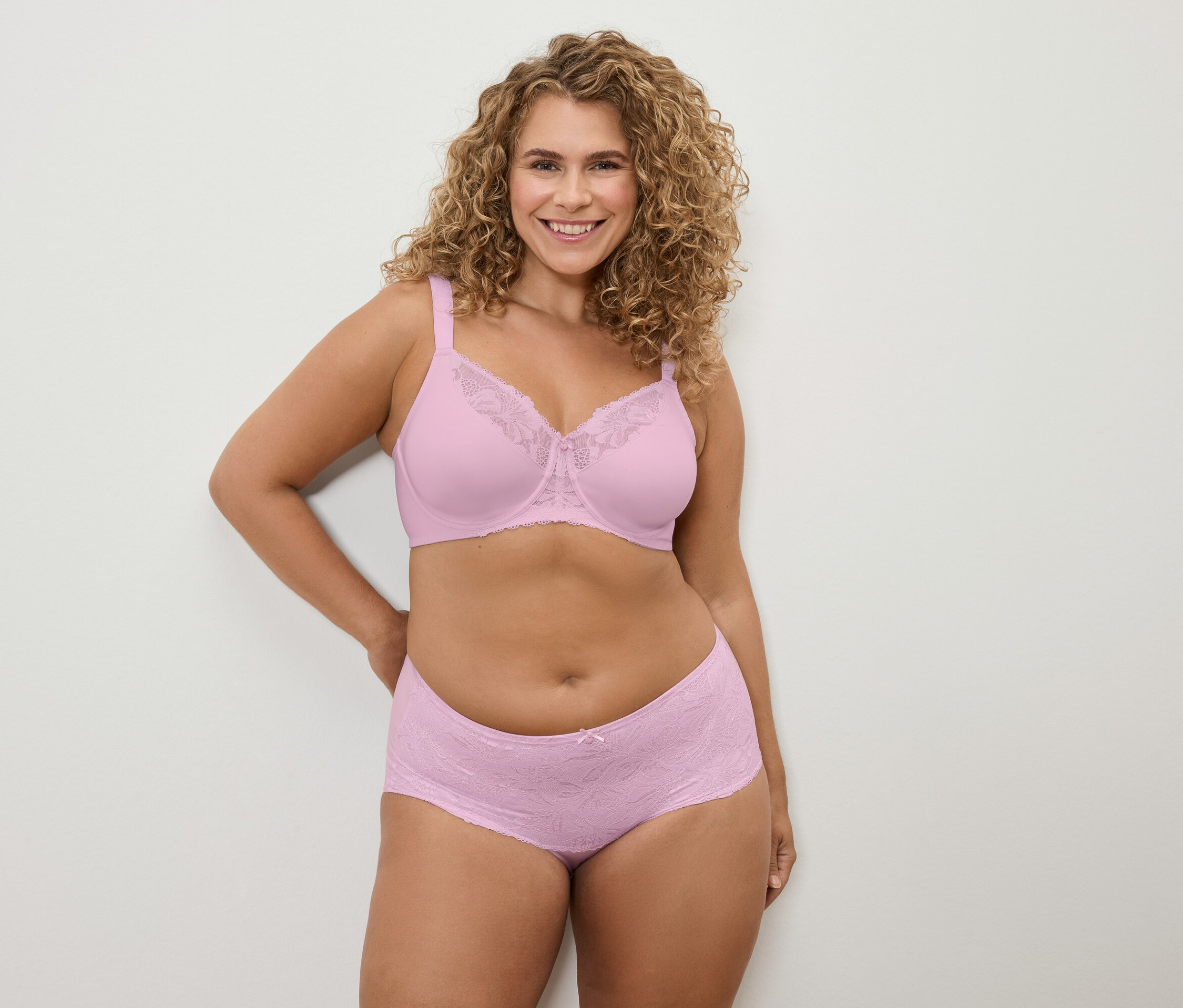 Une femme aux cheveux bouclés pose en soutien-gorge et culotte en dentelle rose devant un mur blanc.