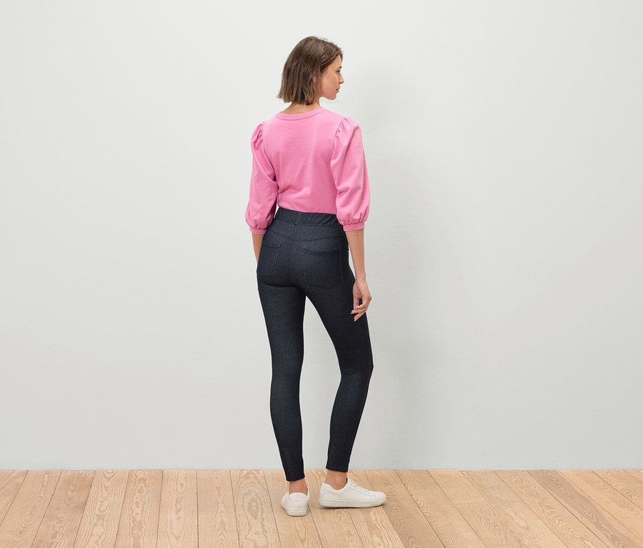 Une femme se tient dos à la caméra, portant un haut rose à manches trois-quarts, un jean foncé et des baskets blanches.