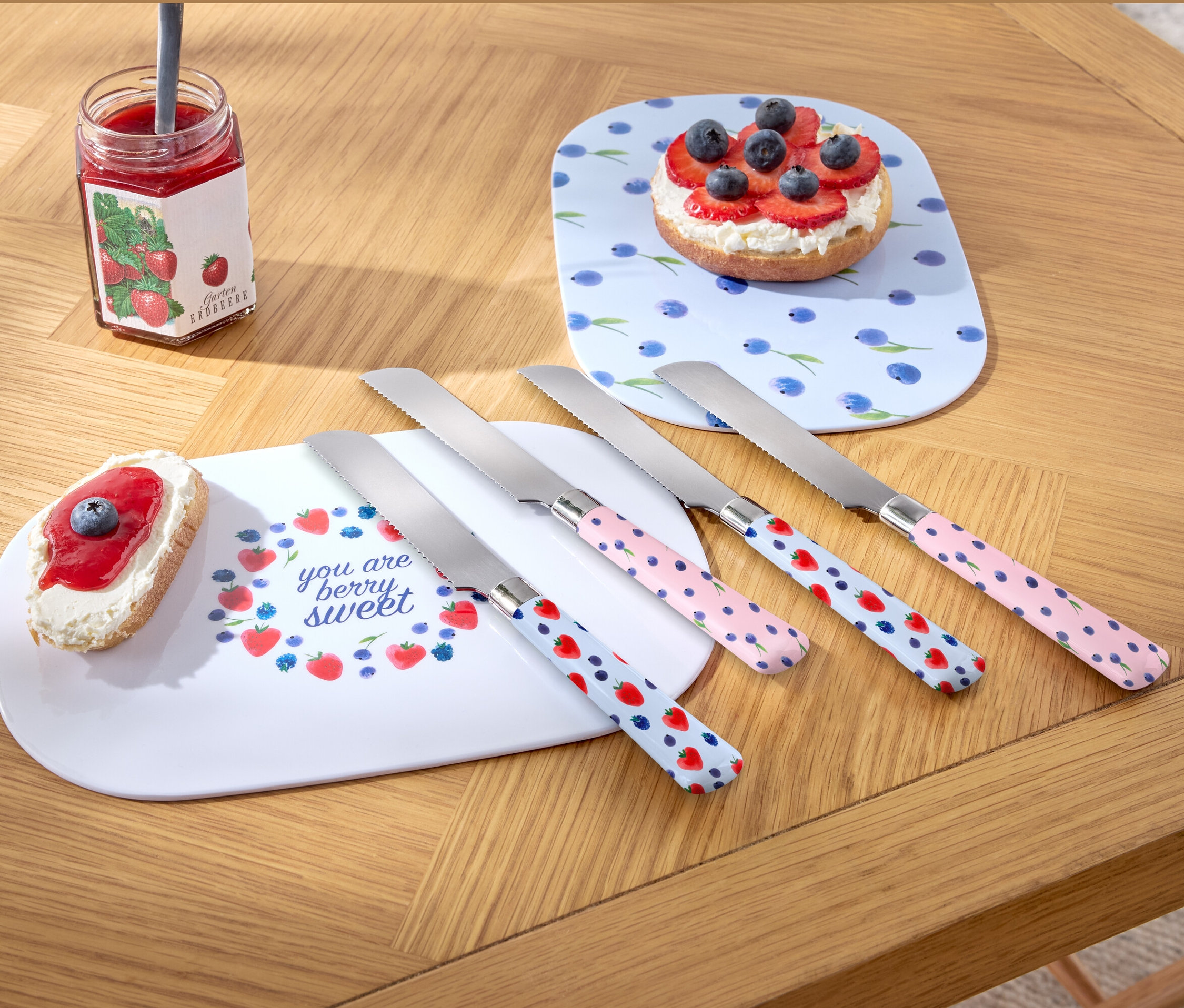 Quatre couteaux à petit-déjeuner sont posés sur une table en bois à côté de deux assiettes et d'un pot de confiture.