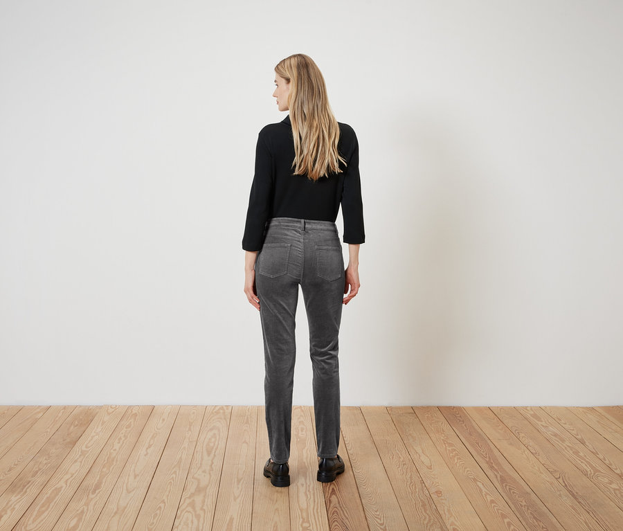 Une femme se tient dos à vous, vêtue d'un chemisier noir et d'un pantalon de velours gris.