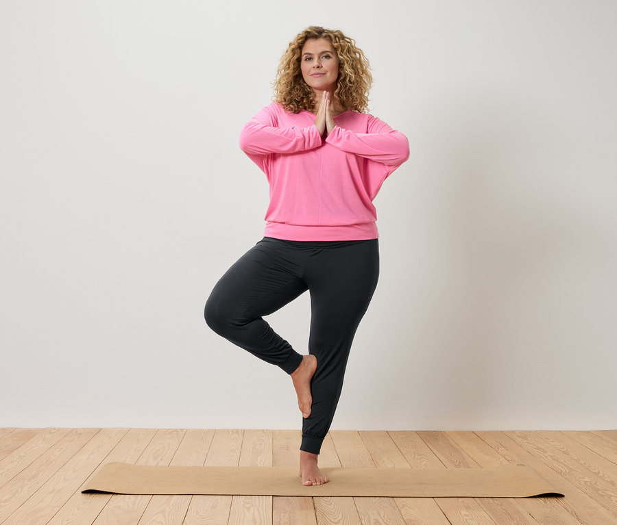 Femme aux cheveux bouclés faisant du yoga sur un tapis beige, vêtue d'un haut rose et d'un pantalon gris.