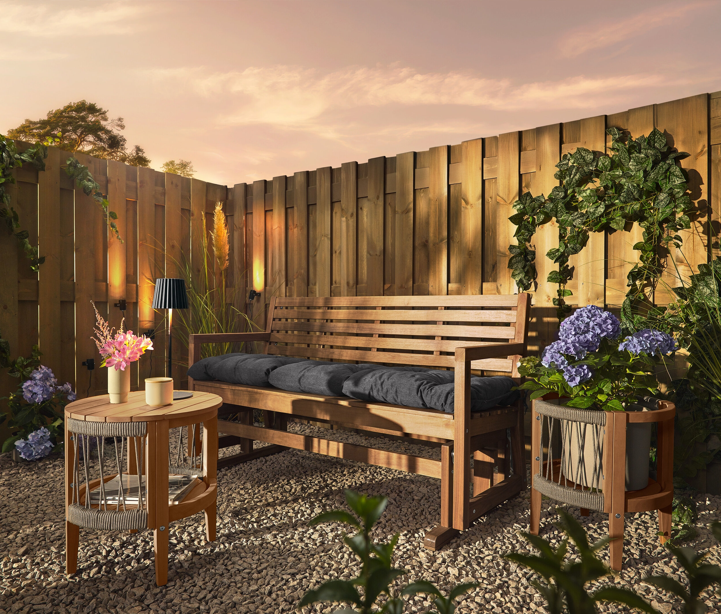 Un banc en bois avec des coussins est placé sur un sol de gravier devant une clôture en bois. Trois lampes solaires pour gouttières sont fixées à la clôture.