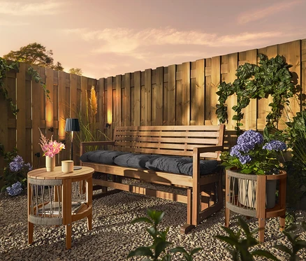 Un banc en bois avec des coussins est placé sur un sol de gravier devant une clôture en bois. Trois lampes solaires pour gouttières sont fixées à la clôture.