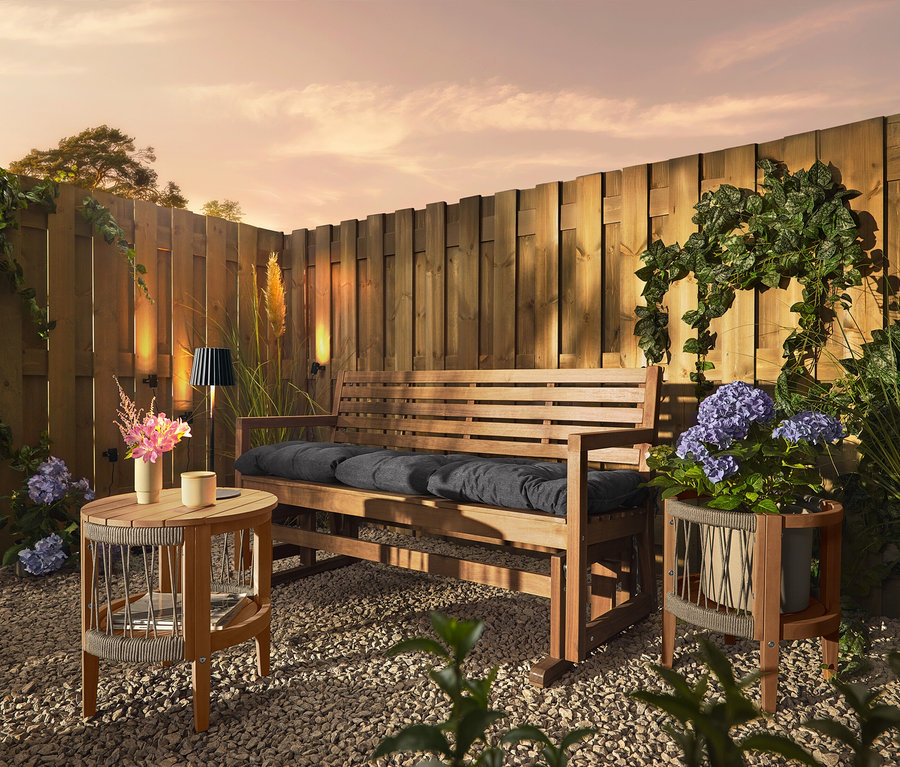 Un banc en bois avec des coussins est placé sur un sol de gravier devant une clôture en bois. Trois lampes solaires pour gouttières sont fixées à la clôture.