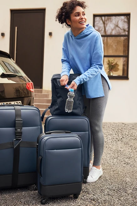 Une femme se tient à côté d'une voiture et ouvre une bouteille d'eau, au premier plan valise hybride petite, moyenne et grande et baskets en cuir.