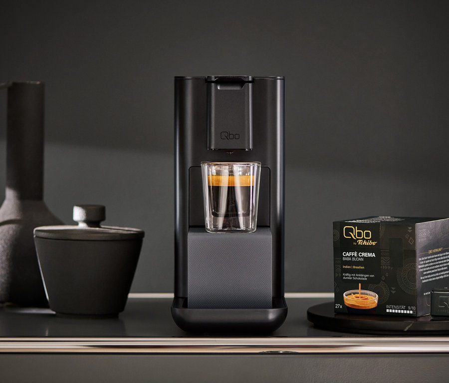 Machine à café Qbo ICONIC Dark Chrome avec tasse en verre et paquet de capsules Qbo sur une table.