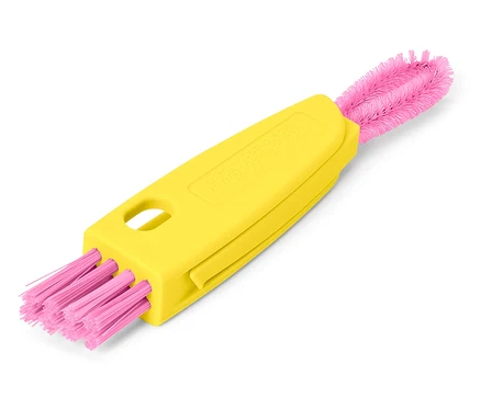 Une brosse de nettoyage jaune à deux extrémités avec des poils roses est posée en diagonale sur un fond blanc.