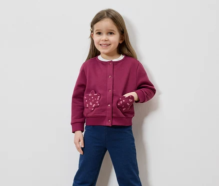 Une fillette souriante aux cheveux bruns porte un cardigan bordeaux avec des poches en forme de fleurs et un pantalon bleu sur fond blanc.