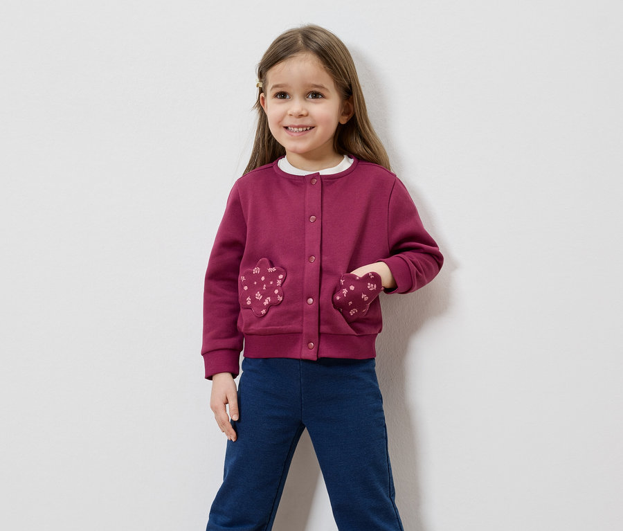 Une fillette souriante aux cheveux bruns porte un cardigan bordeaux avec des poches en forme de fleurs et un pantalon bleu sur fond blanc.