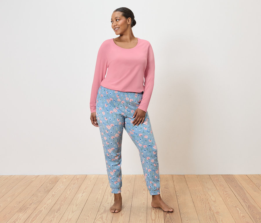 Femme debout pieds nus, portant un pull rose et un pantalon bleu clair à motifs floraux.