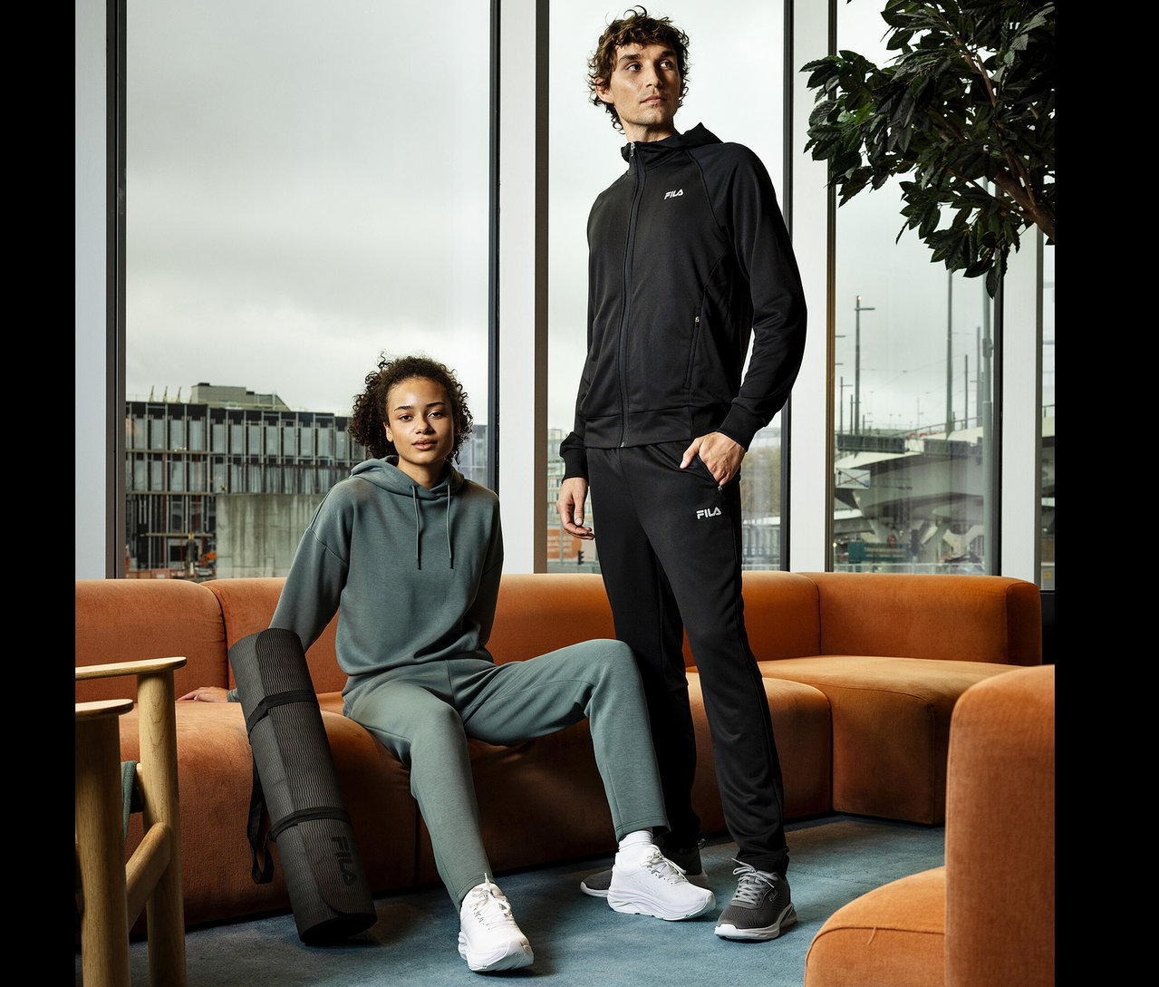 Une jeune femme est assise sur un canapé orange et tient un tapis de yoga noir. Un jeune homme en survêtement noir se tient à côté d'elle.