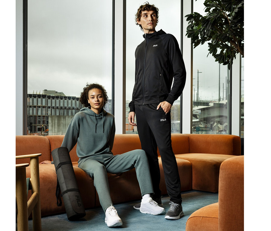 Un homme portant une veste sweat technique pour homme FILA noire se tient à côté d'une femme en sweat à capuche et pantalon de jogging verts, assise avec un tapis de yoga sur un canapé orange.