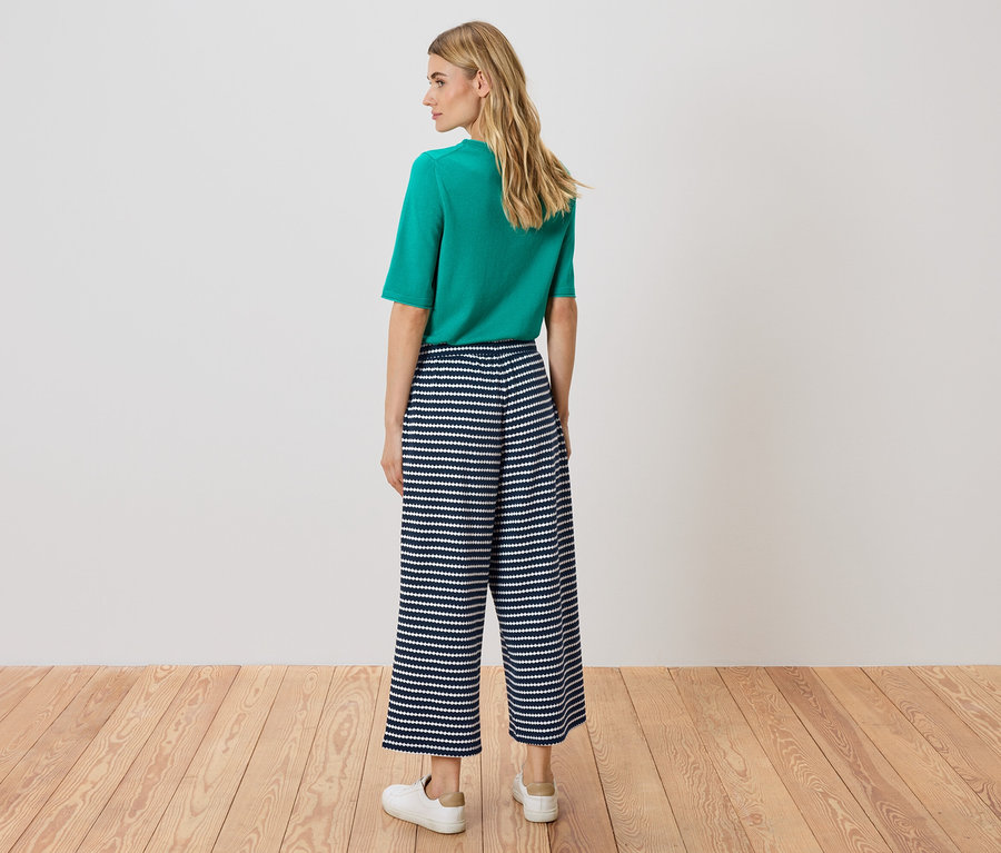 Une femme se tient dos à la caméra, portant un haut vert et un pantalon culotte rayé bleu et blanc.