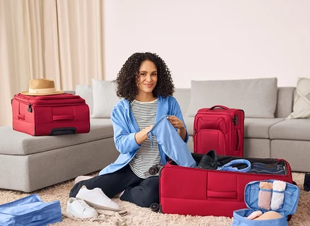Une femme est assise par terre et range un pantalon de jogging bleu foncé avec des poches cargo dans une valise rouge. À côté d'elle se trouvent des baskets en cuir.