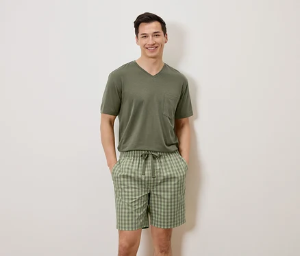 Un homme se tient devant un mur blanc, les mains dans les poches, portant un t-shirt vert olive et un short à carreaux.