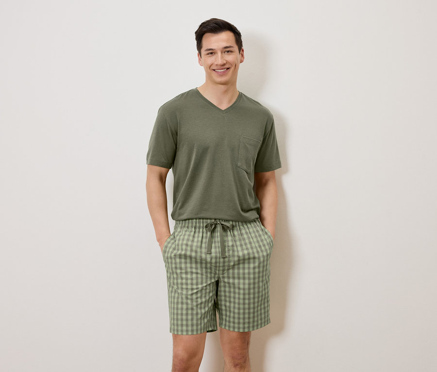 Un homme se tient devant un mur blanc, les mains dans les poches, portant un t-shirt vert olive et un short à carreaux.