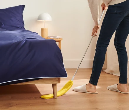 Une femme nettoie avec un plumeau jaune sous un lit avec des draps bleu marine.