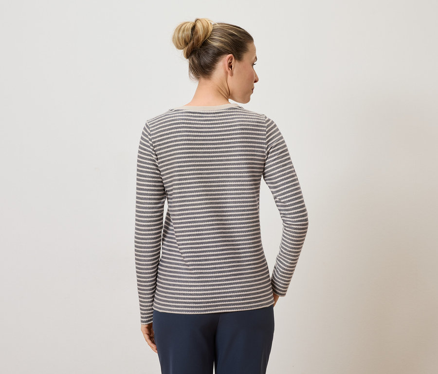 Une femme portant un chignon, vue de dos, porte un t-shirt à manches longues rayé gris et blanc et un pantalon bleu sur un fond clair.