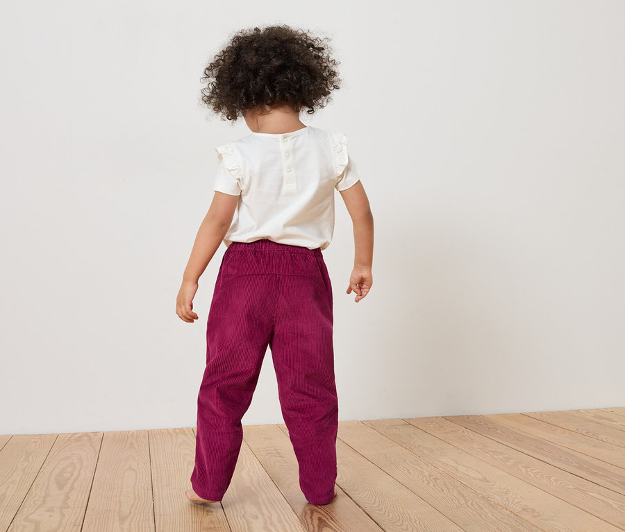 Vue arrière d'un enfant aux cheveux bouclés portant un t-shirt blanc et un pantalon en velours côtelé bordeaux sur un plancher en bois.