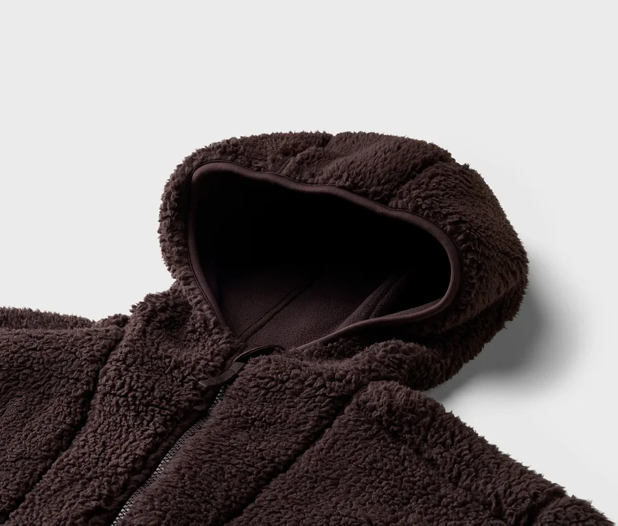 Gros plan d'une veste marron en tissu peluche avec capuche et fermeture éclair sur fond gris clair.