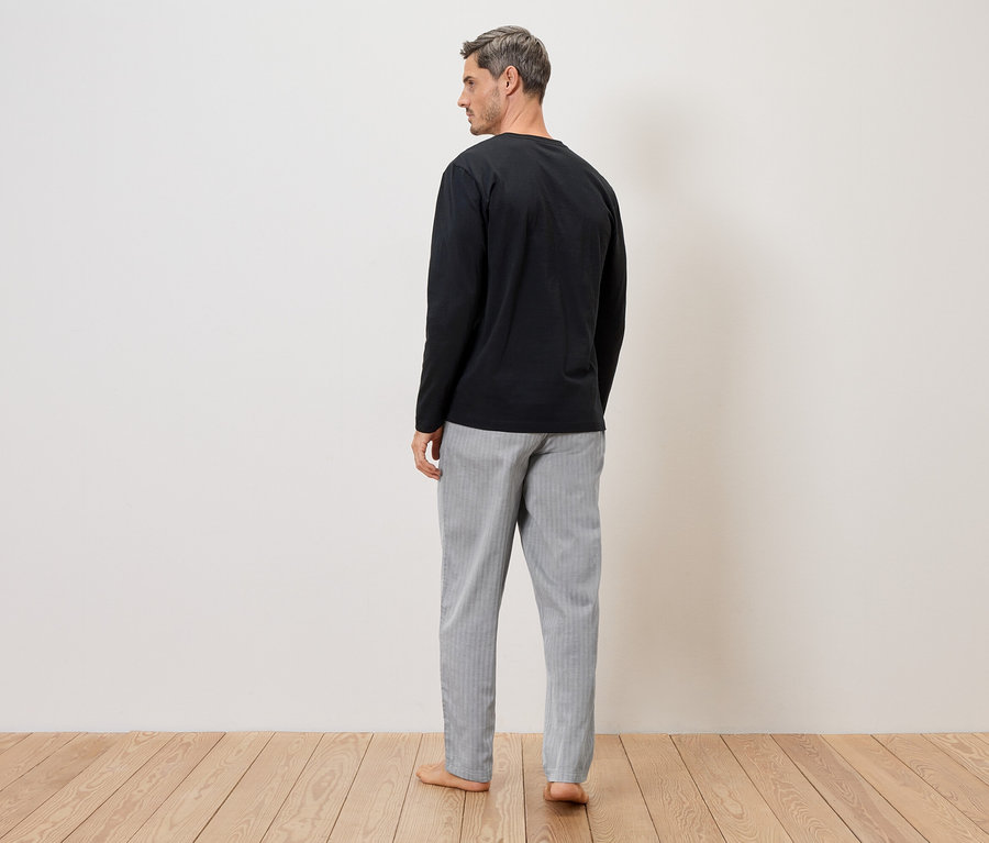 Homme vu de dos portant un t-shirt noir à manches longues et un pantalon rayé gris et blanc devant un mur blanc sur un plancher en bois.