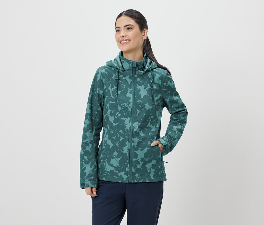 Une femme souriante portant une veste à capuche verte à motif floral et un pantalon bleu marine sur fond blanc.