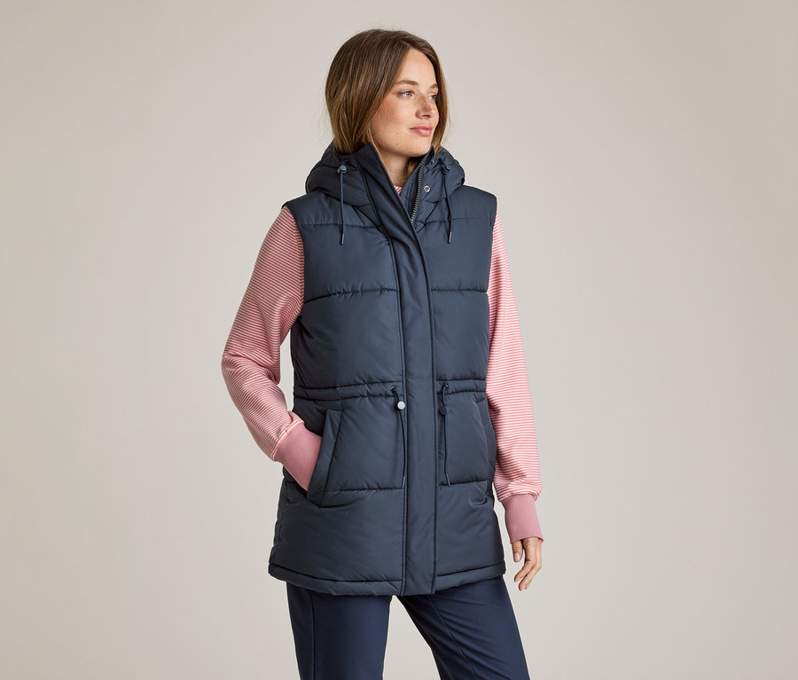 Femme portant un gilet doudoune à capuche bleu marine sur un t-shirt à manches longues rayé rose et blanc et un pantalon bleu marine, la main gauche dans la poche, regardant vers la droite.