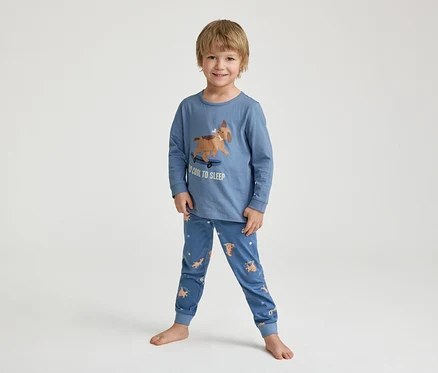 Un petit garçon souriant est debout pieds nus, vêtu d'un pyjama bleu avec un imprimé de chien sur un skateboard et l'inscription "TOO COOL TO SLEEP".