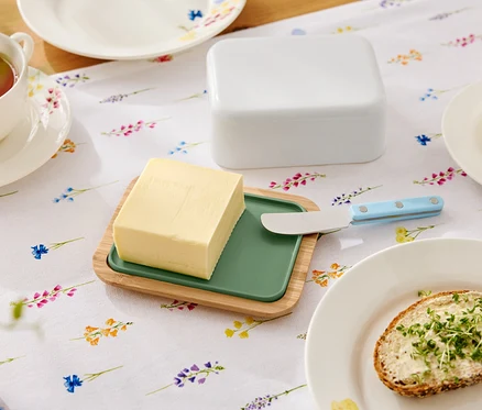 Sur une table fleurie se trouvent du beurre sur une assiette en bois avec un insert vert, un couteau à beurre, un beurrier, une tasse de thé et une assiette avec une tartine beurrée.
