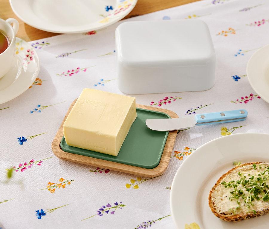 Sur une table fleurie se trouvent du beurre sur une assiette en bois avec un insert vert, un couteau à beurre, un beurrier, une tasse de thé et une assiette avec une tartine beurrée.