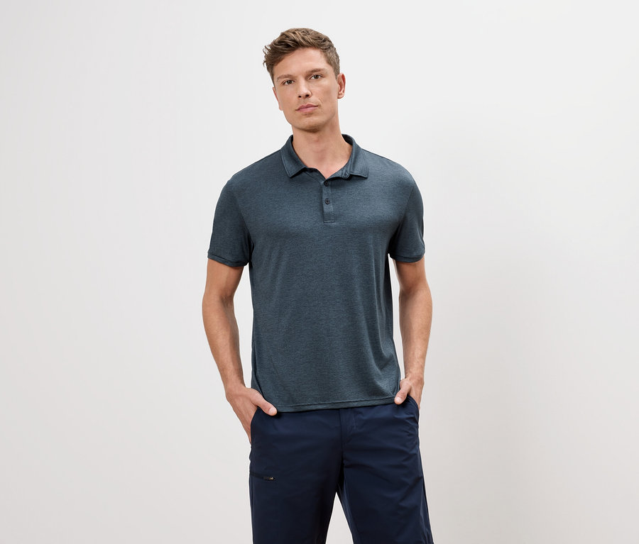 Un homme portant un polo bleu et un short bleu se tient devant un fond blanc.