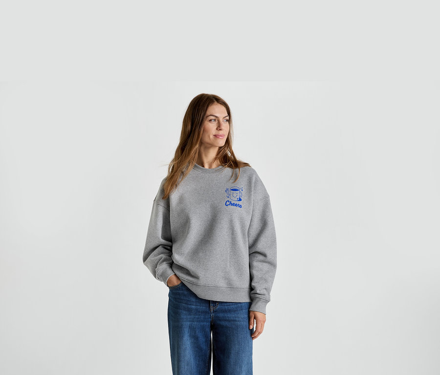 Femme portant un sweat gris avec un imprimé bleu 'Cheers' et un jean.