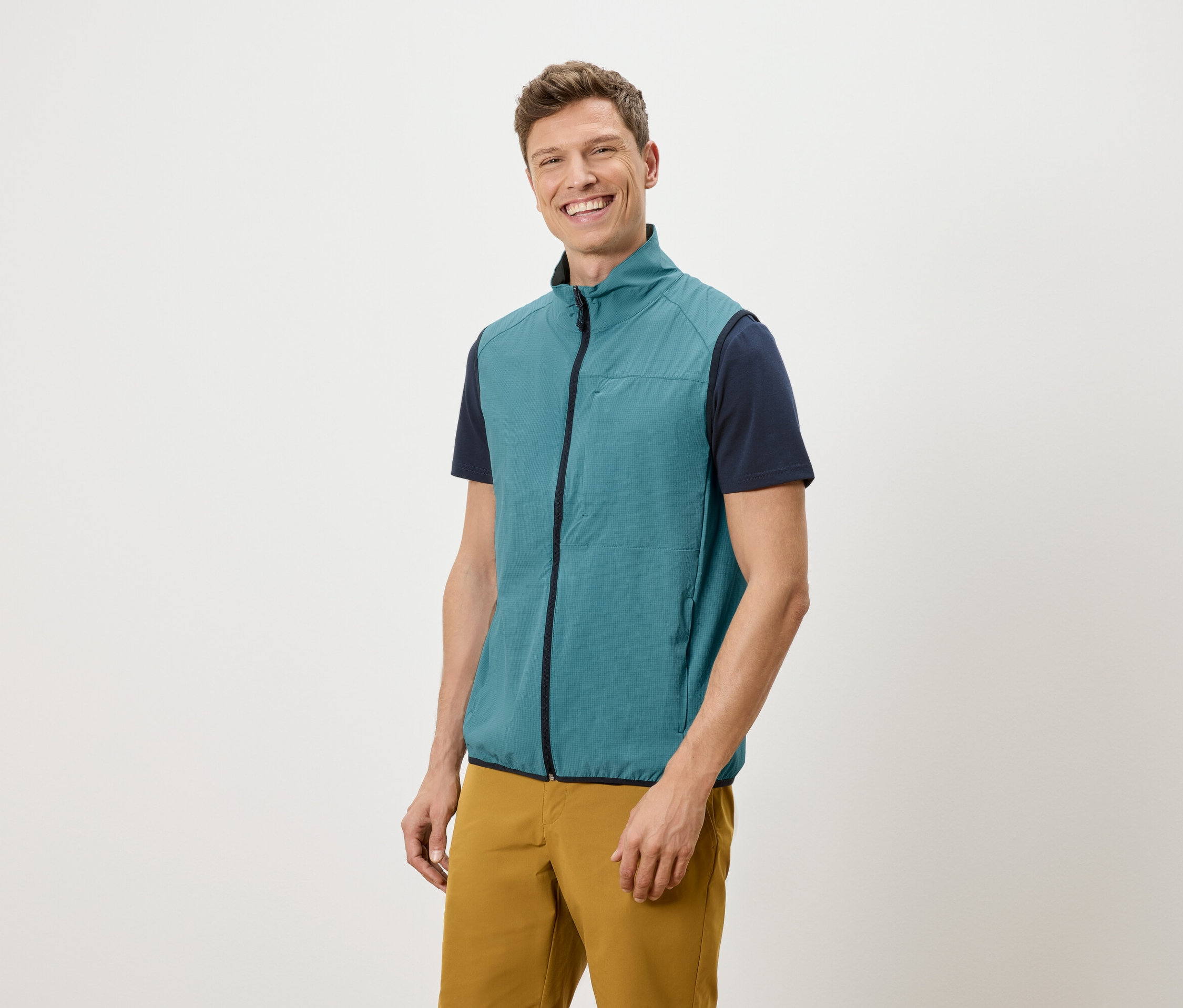 Un homme souriant portant un gilet bleu pétrole, un t-shirt bleu marine et un pantalon ocre se tient devant un fond blanc.