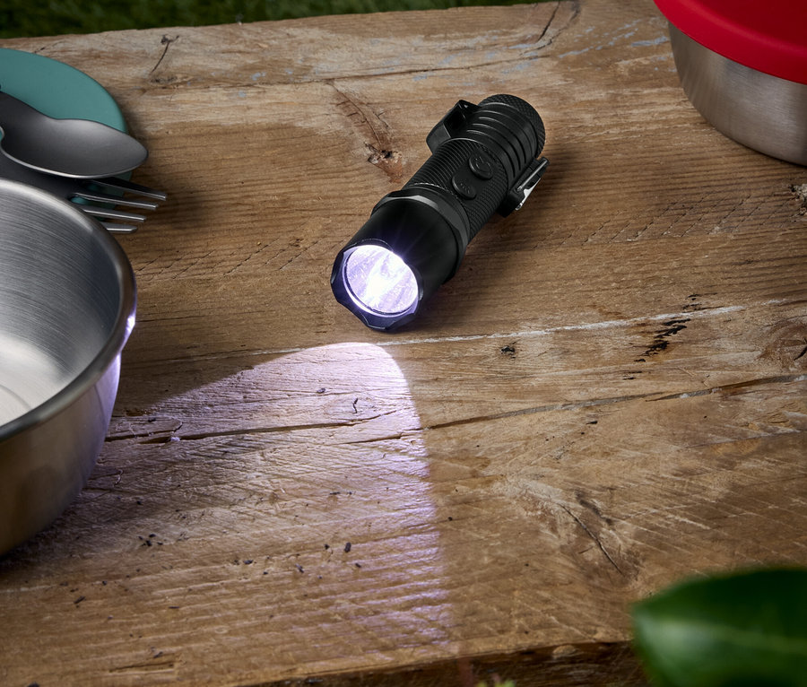 Une lampe de poche noire brille sur une table en bois, à côté d'un bol, d'assiettes et d'un récipient avec un couvercle rouge.