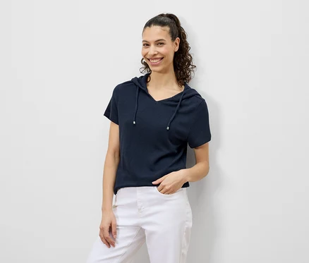 Une femme aux cheveux bouclés sourit dans un t-shirt à capuche bleu marine et un pantalon blanc.