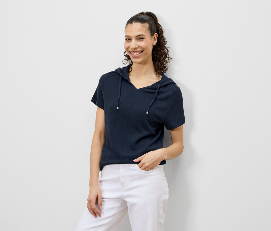 Une femme aux cheveux bouclés sourit dans un t-shirt à capuche bleu marine et un pantalon blanc.