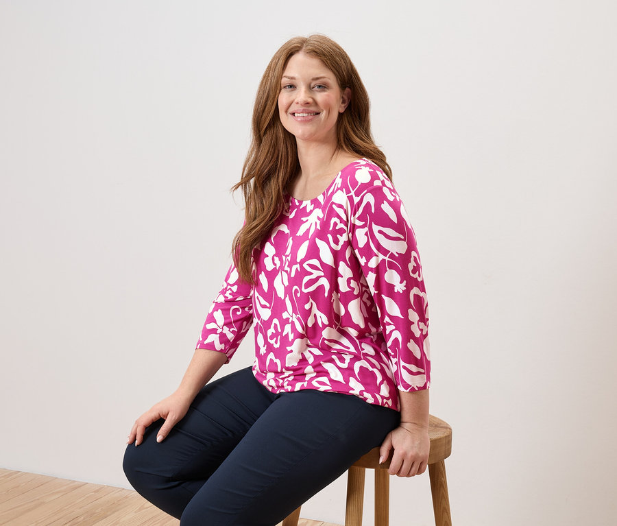 Une femme aux longs cheveux roux est assise sur une chaise en bois, portant un chemisier rose à motifs de feuilles blanches et un pantalon bleu marine.