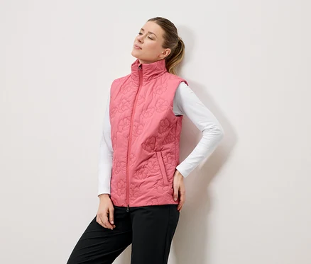 Une femme pose devant un mur blanc, vêtue d'un gilet rose matelassé à motifs floraux, d'un chemisier blanc à manches longues et d'un pantalon noir.