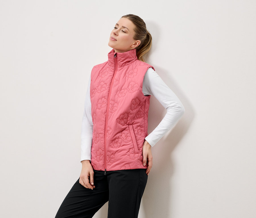 Une femme pose devant un mur blanc, vêtue d'un gilet rose matelassé à motifs floraux, d'un chemisier blanc à manches longues et d'un pantalon noir.