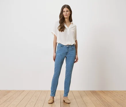 Une femme en jean bleu clair, chemisier beige et chaussures se tient devant un mur blanc sur un plancher en bois.