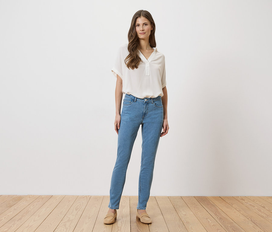 Une femme en jean bleu clair, chemisier beige et chaussures se tient devant un mur blanc sur un plancher en bois.