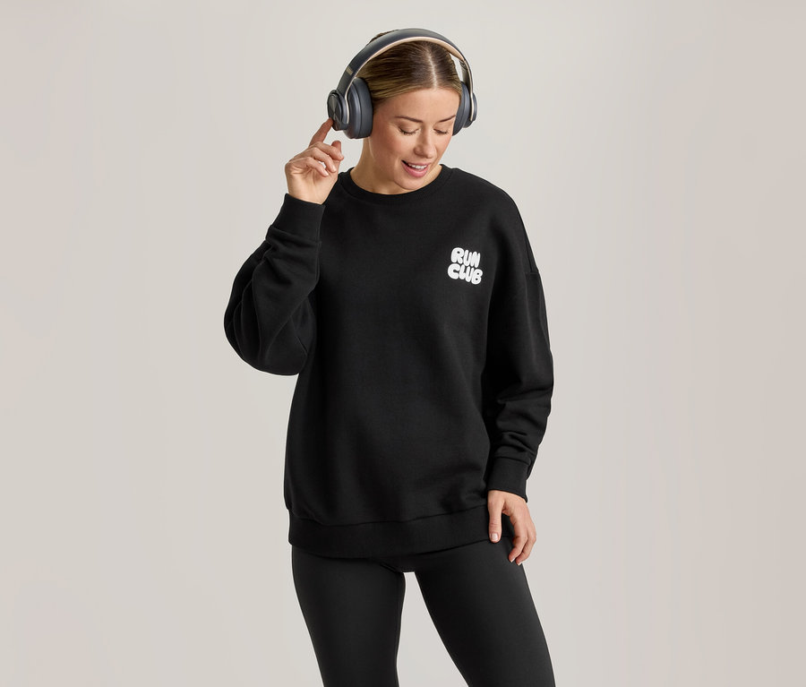 Une femme portant un sweatshirt noir avec l'inscription « RUN CLUB », un legging noir et des écouteurs gris, souriant et touchant son écouteur droit.