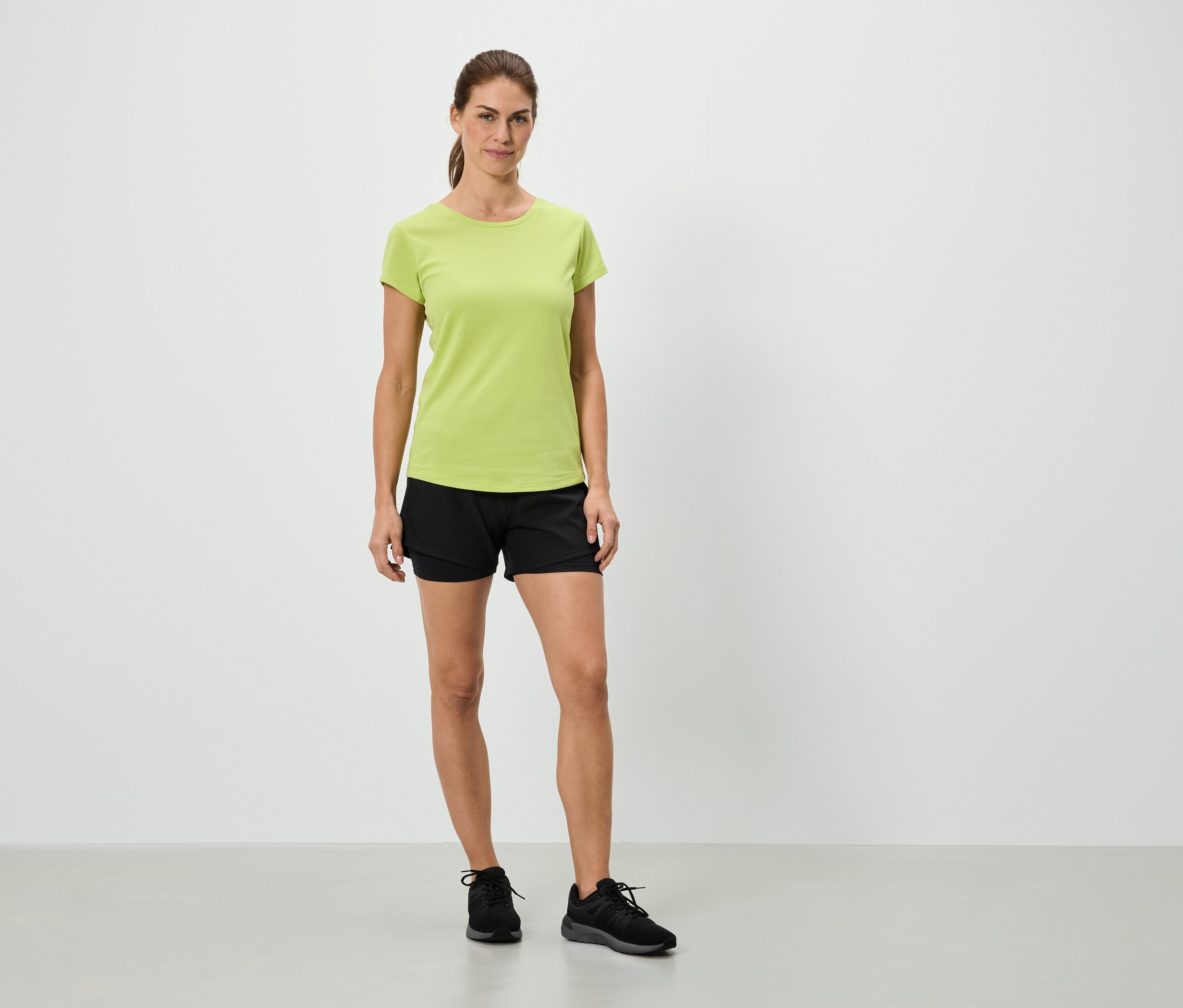 Une femme se tient debout devant un mur blanc, portant un t-shirt vert clair, un short noir et des baskets noires.