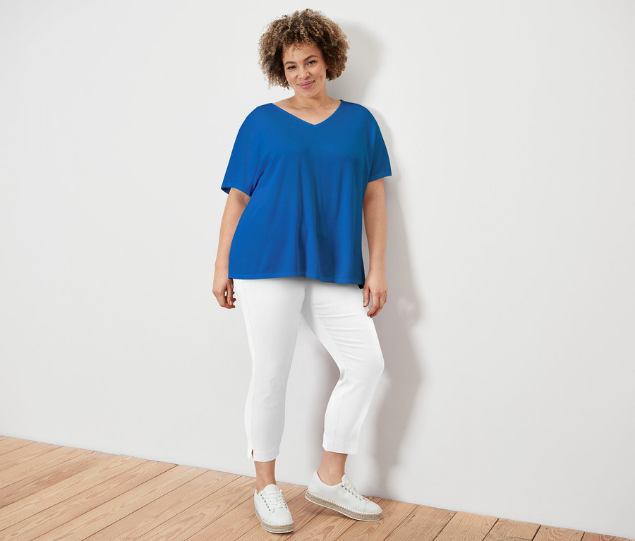 Une femme aux cheveux bouclés porte un t-shirt bleu à col en V, un pantalon blanc et des chaussures blanches, debout contre un mur blanc.