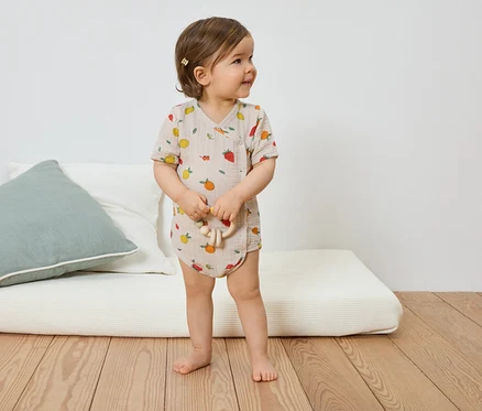 Un jeune enfant avec une barrette porte un body à manches courtes marron clair à motifs de fruits et tient un jouet en bois.
