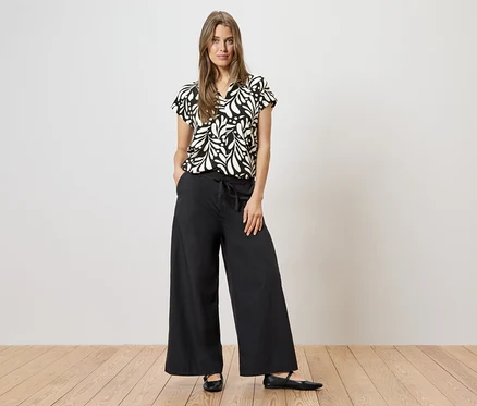 Une femme portant un chemisier à motifs noirs et blancs et un pantalon noir à jambes larges, debout sur un plancher en bois.