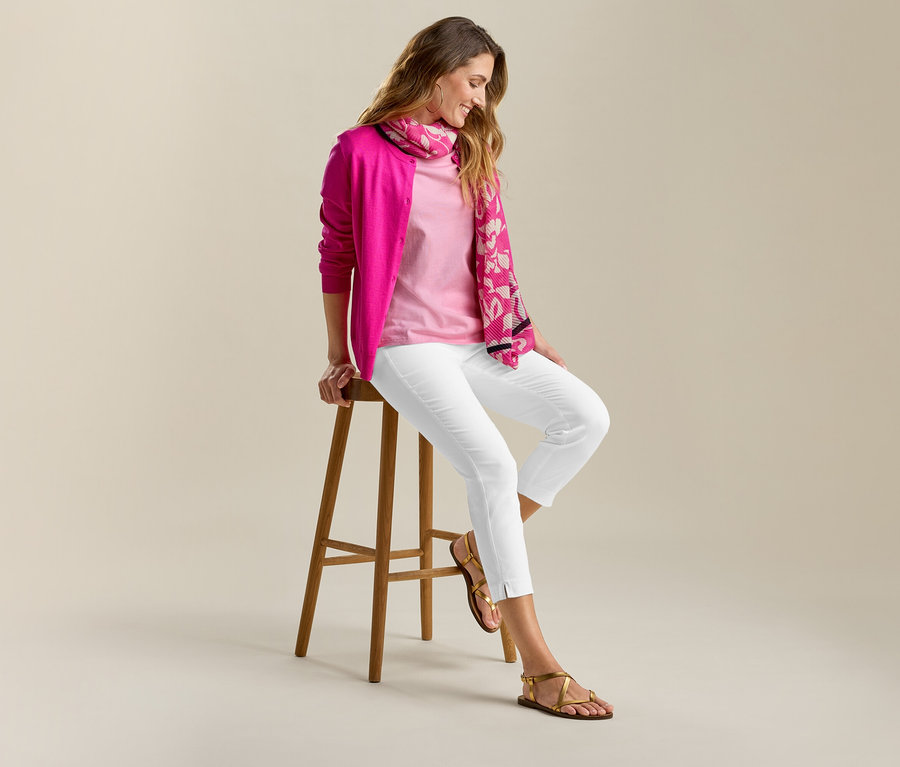 Femme assise sur un tabouret avec un cardigan en maille fine et un pantalon stretch 7/8 blanc. Elle porte des sandales et une écharpe.