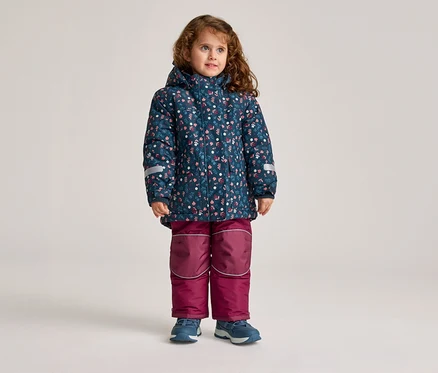 Une fillette aux cheveux bouclés se tient debout, portant une veste bleue à fleurs et un pantalon bordeaux.
